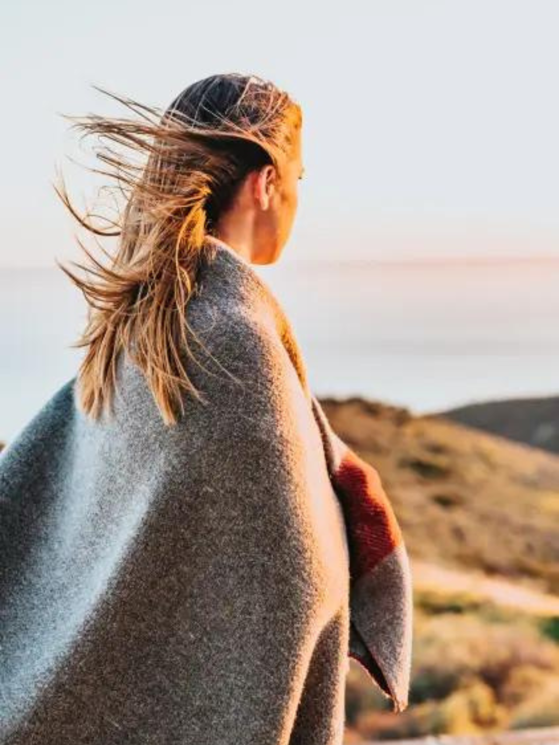 A woman stares off into the sandy distance wearing a grey wool blanket wrapped around her shoulders. Her hair is windblown.