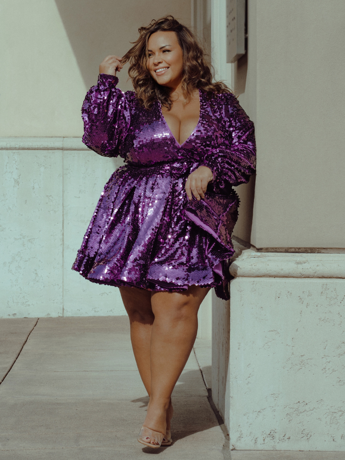 A woman leans against a wall, wearing a short purple sparkly dress.
