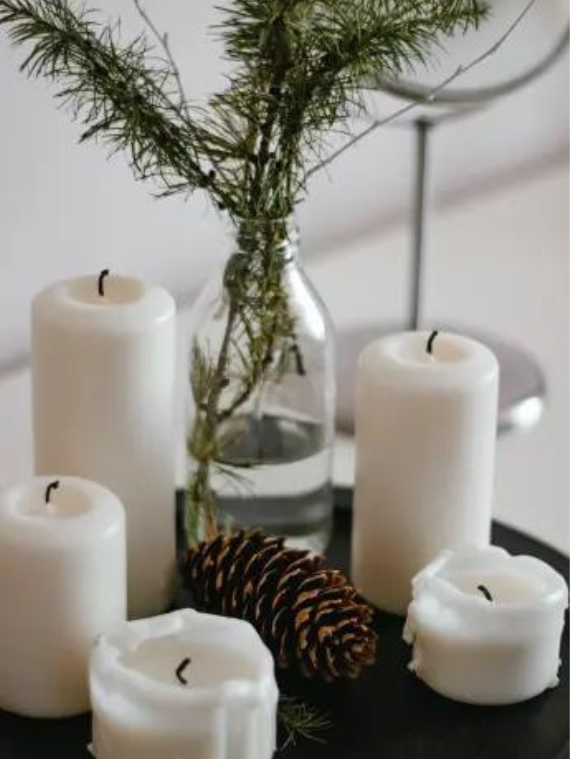 White pillar candles sit on a table. A vase holds a fresh pine or fir sprig.